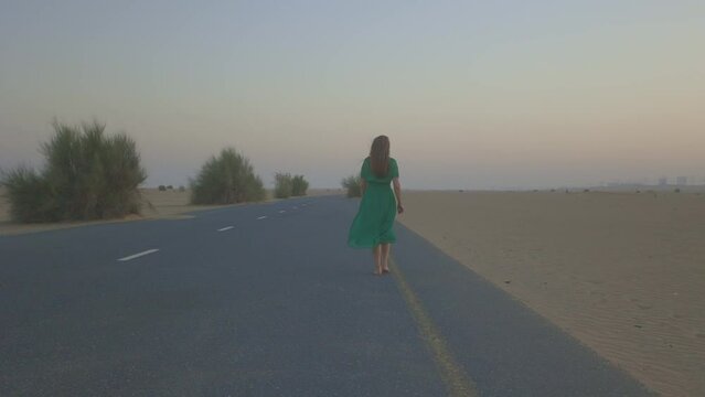 A beautiful young woman in green dress walks barefoot along road in the desert next to a car.  black car stops at the girl's door. A man invites beautiful girl into car