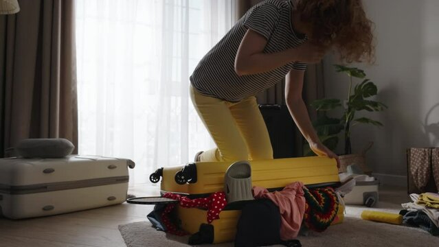 Woman try to close travel luggage, sits down with knees on suitcase. Woman in hurry panic tries to pack luggage, close suitcase. Impulsive unplanned trip travel, late for airport flight. Slow motion.
