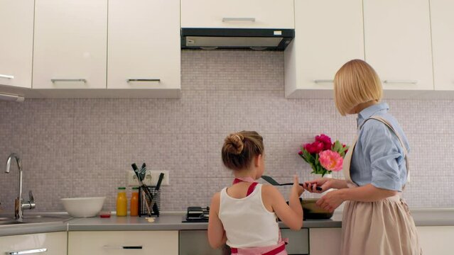 A young mother and her little daughter cook pancakes together for breakfast. Mother teaches daughter to cook.