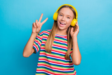Portrait of good mood positive teen girl wear colorful t-shirt in headphones showing v-sign symbol isolated on blue color background