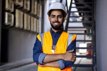 Portrait of young successful engineer worker, man smiling and looking at camera, wearing hard hat and vest, working in factory plant.