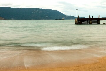 Praia de areia vermelha coo movimento das ondas borrado e píer de madeira ao fundo. Praias de Ilha Bela, Litoral Norte de São Paulo, Brasil. 