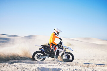 Extreme sport, motorcycle and athlete with sand by wheels with pride, skill and adrenaline rush. Biker, sunshine and dust with blue sky in fun, fitness and achievement in speed, race and lens flare