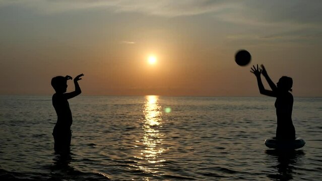 Two teenagers play ball while swimming in the ocean at sunset. A boy and a girl throw a ball to each other while standing in the sunset sea. Scenic slow motion footage. Slow motion