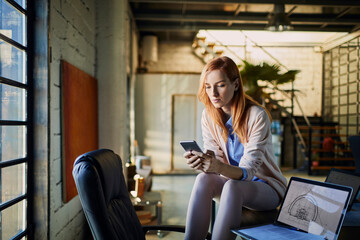 Fototapeta premium Young Caucasian female designer using her smartphone while working in startup company office