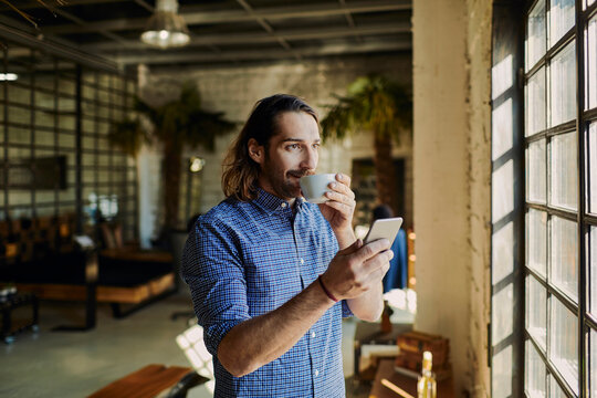 Young Caucasian male designer enjoying a cup of coffee and using his smartphone while on a break in a startup company office