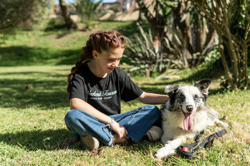 Red haired girl with braids sitting on the grass in a park playing with her black and white dog.