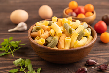 Rigatoni colored raw pasta with tomato, eggs, spices, herbs on brown wooden. Side view, selective focus