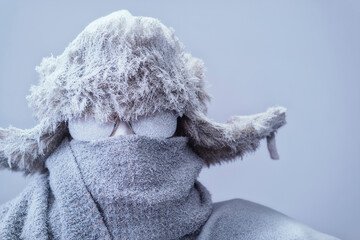 A frozen man all bundled up in a fur trappers hat, scarf, and parka, covered in snow and frost trying to stay warm on a very cold gray Winter's day.