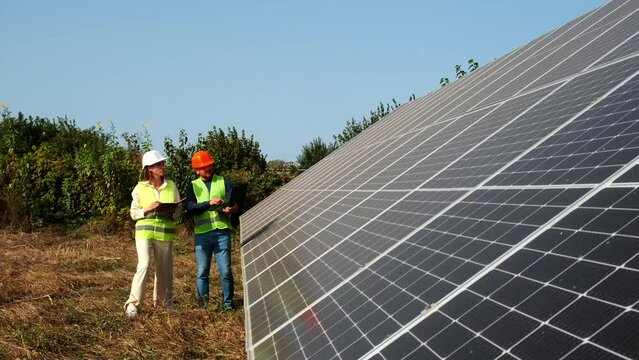 A man and a woman in hard hats and with a laptop are discussing work tasks against the backdrop of solar panels outside. Female environmental engineer talking to investor. Green electricity concept.