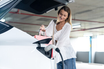Young woman travel with EV electric car to shopping center parking lot charging in downtown city...