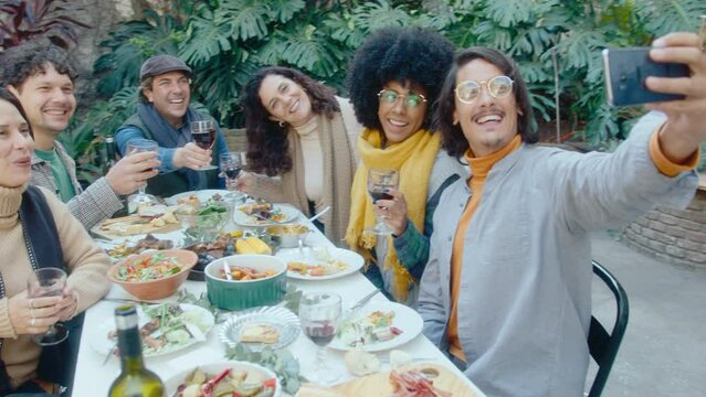 Joyous man holding smartphone and taking selfies with smiling friends sitting around dinner table with food and drinks on outdoor party