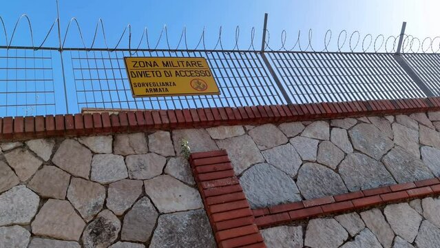 A sign attached to a fence with barbed wire with the words military zone no access to armed surveillance