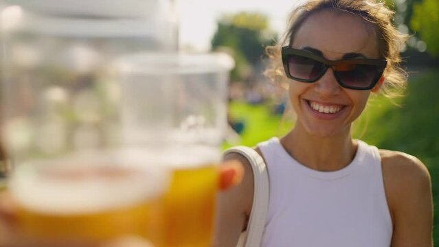 POV happy joyful young woman have fun at outdoors picnic on green meadow. woman holding plastic glass with beer and cheers with someone. party time outdoors. Happy hour, lunch break and youth concept