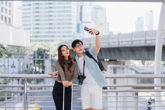 Asian tourist couple standing with luggage before going on summer vacation or honeymoon trip in airport or bus station or train station. Couple take photos together, happy moment traveling in the city
