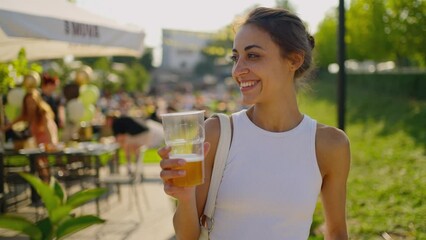 POV Portrait of beautiful happy young woman drinking beer and enjoying picnic at bravery beerpub backyard summer day. woman holding plastic glass with beer. party time outdoors.