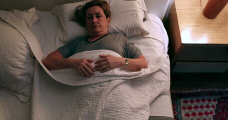 Older woman prepares to go to bed and sleep. Person lays down in bed and turns off night stand besides bed