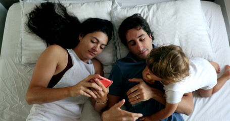 Millennial couple in bed with infant toddler boy wanting attention. Man and woman lying down looking at cellphone, top view