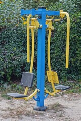 one sports modern iron athletic new blue and yellow public industrial for pumping muscles and fitness machine stands on the ground on the sports ground near the green bushes