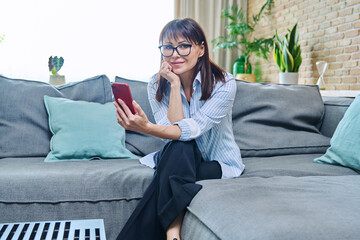 Middle-aged woman with smartphone sitting on sofa in living room interior