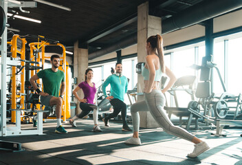 Group of people exercising in a health club. Cheerful young athletes doing lunges in gym under...