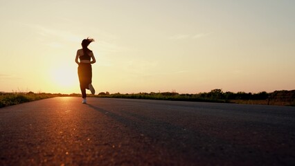 Running after sun. Training jogging. Beautiful girl doing fitness, jogging on road in sun. Jogger girl breathes fresh air in nature. Free young woman runs in summer park, sunset. Jogging outside city