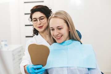 Fototapeta premium Young dentist and her young female patient smiling while looking in the mirror in dental clinic