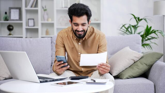 Happy man uses mobile app for spending bills using smartphone on phone while sitting at home in room. A male calculates household finances and enters data into a chat bot easy and simple to pay