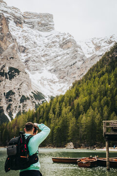 Fot&oacute;grafo con mochila, captando momentos con la c&aacute;mara en lago con monta&ntilde;as nevadas y estructura de madera. 