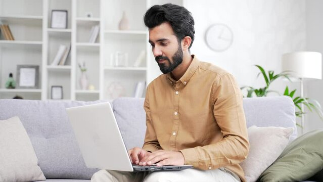 Confident Young Bearded Male Freelancer Typing On Laptop While Sitting On Sofa In Living Room At Home Office. A Serious Developer, Coder Or IT Programmer Works On A Computer Remotely On A Project