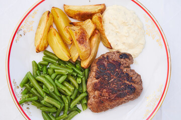 Close up picture on the delicious beef steak from organic farming served with boiled green beans, wedges of baked potatoes and tartar sauce on the rustic style porcelain plate. Balanced healthy dish.