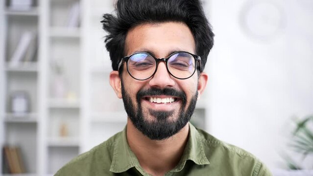 Close up portrait of young handsome bearded businessman in shirt sitting in living room at home office. Head shot of positive coder in glasses. Smiling IT programmer developer poses looking at camera