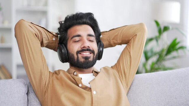 Calm young handsome bearded man in wireless headphones listening to relaxing music while sitting on sofa in living room at home. Happy male enjoys to the sounds of relaxation and the noise of nature