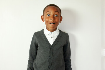 Amazed, shocked, astonished black school kid in cardigan standing against white studio background with big round eyes and surprised overjoyed facial expression after winning abroad study grant