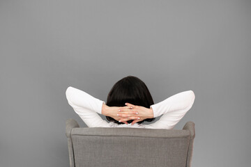 Rear view of unrecognizable woman with hands behind head sits on comfortable chair isolated on grey...