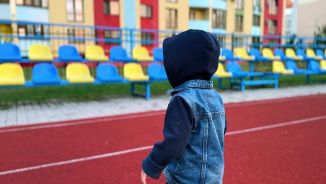 Cute Caucasian toddler in hoodie and jeans jacket walks by the stadium. Kid goes to the spectators' seat in two rows.