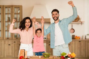 Joyful family holding hands and shaking fists, gesturing yes while preparing dinner together in...