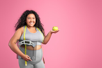 Glad black body positive woman in sportswear holding apple and scales, enjoying diet result, pink background, copy space