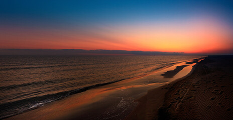 Sunset panorama with orange sky and reflection on the beach and water. Mediterranean, Camargue, Gard, France.