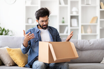 Shocked Indian Man Opening Delivery Box While Sitting On Couch At Home