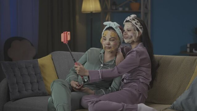 Medium-full video capturing two young women in pajamas sitting on the couch and doing their evening skincare routine, taking photos using a selfie stick, wearing face masks.