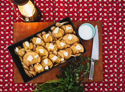 scalloped potatoes patted with butter in a steel pan on a wood cutting board. Nearby are herbs, salt, a knife, and a rustic gas lantern, all atop a red patterned tablecloth
