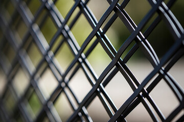 Closeup of an iron fence with a lattice design, featuring thin metal bars crisscrossing to create a woven pattern. The black metal has a matte finish, giving the fence a subtle yet striking