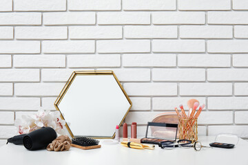 Different makeup products, hair dryer, mirror and beautiful flowers on dressing table near light brick wall in room