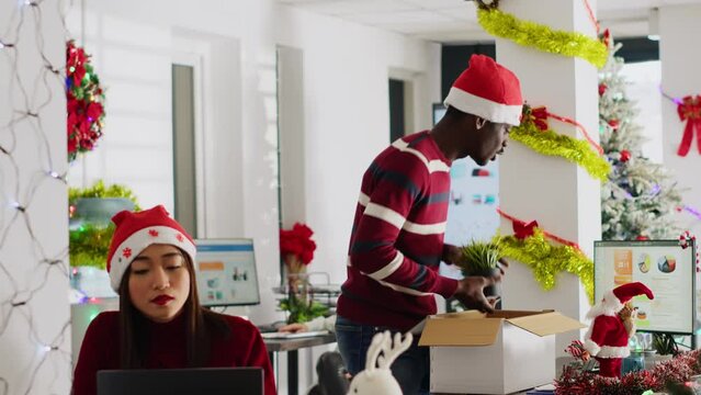Exicted african american employee clearing desk before leaving for vacation, dancing around festive decorated office. Man wearing Santa hat taking days off to go on xmas holiday, feeling merry