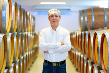 Portrait of Asian man winemaker working and wine quality control in wine cellar with wooden barrels at wine factory. Winery manufacturing industry, brewery, alcohol liquor shop and winemaking concept.