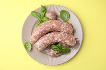 Raw homemade sausages and basil leaves on yellow background, top view
