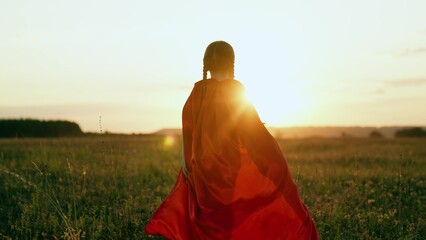 Little girl with long red cloak walks across sunset meadow superhero party