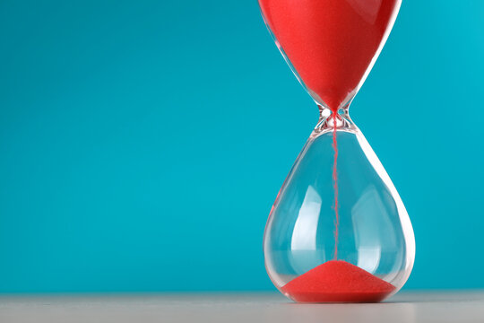 Hourglass with flowing red sand on table against light blue background, space for text
