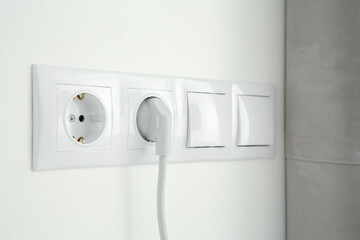 Power sockets with inserted plug and light switches on white wall indoors